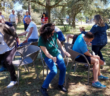Personnel from Hexagon and the Gil Gayarre Foundation play musical chairs. They are outside in grass, near a tree